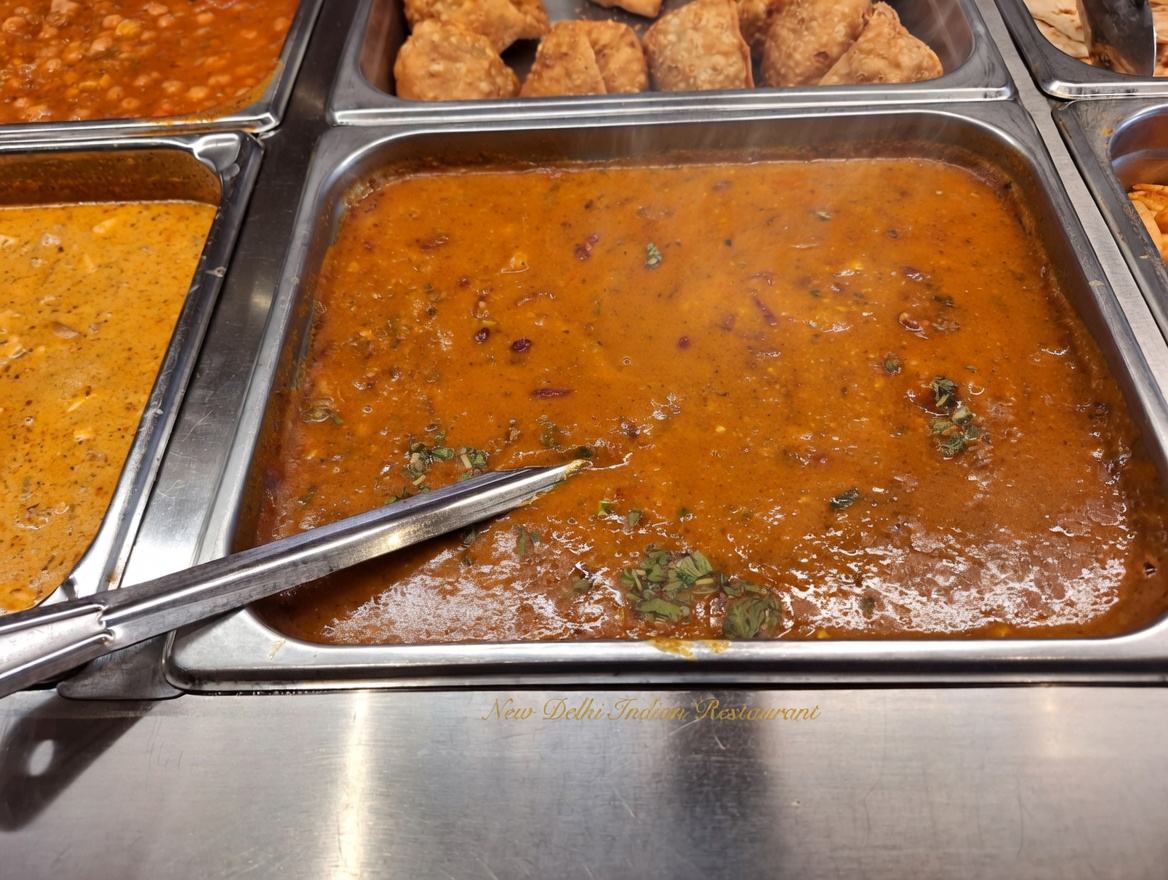 Close-up of a rich, aromatic orange curry simmering in a stainless steel steam table pan at the New Delhi Indian Restaurant buffet, with metal serving tongs
