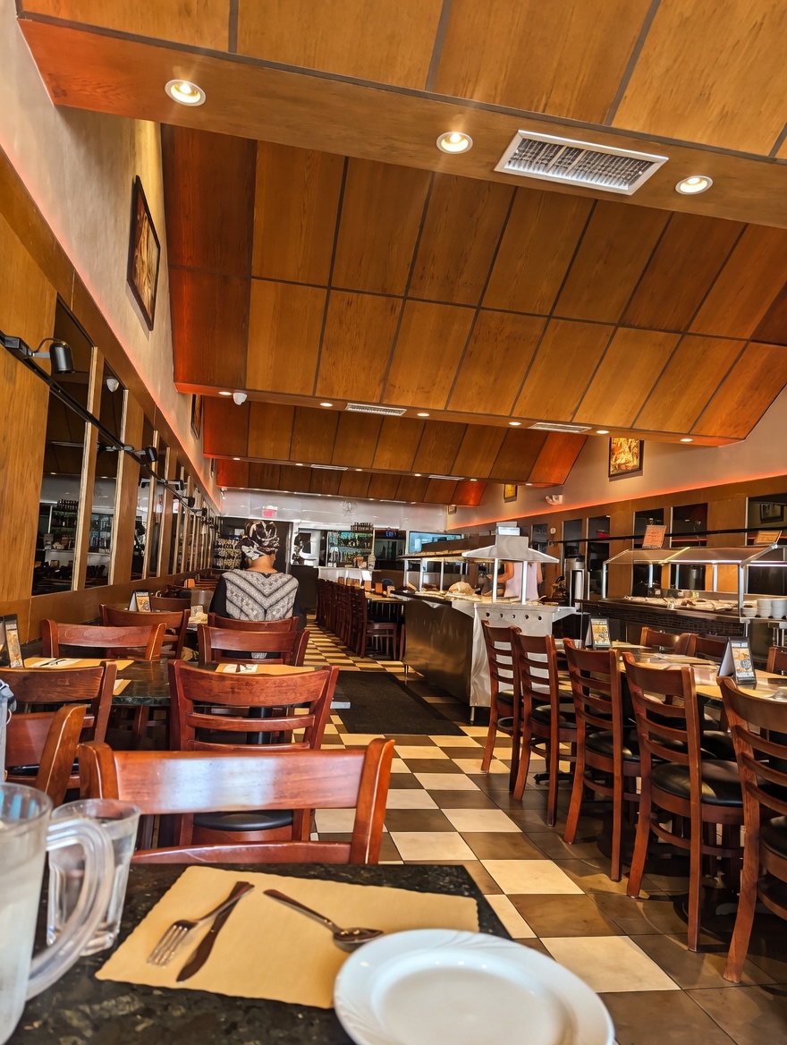 Interior view of the dining area at New Delhi Indian Restaurant looking toward the back, with tables set for service, a guest seated, and the stainless steel buffet line visible in the background
