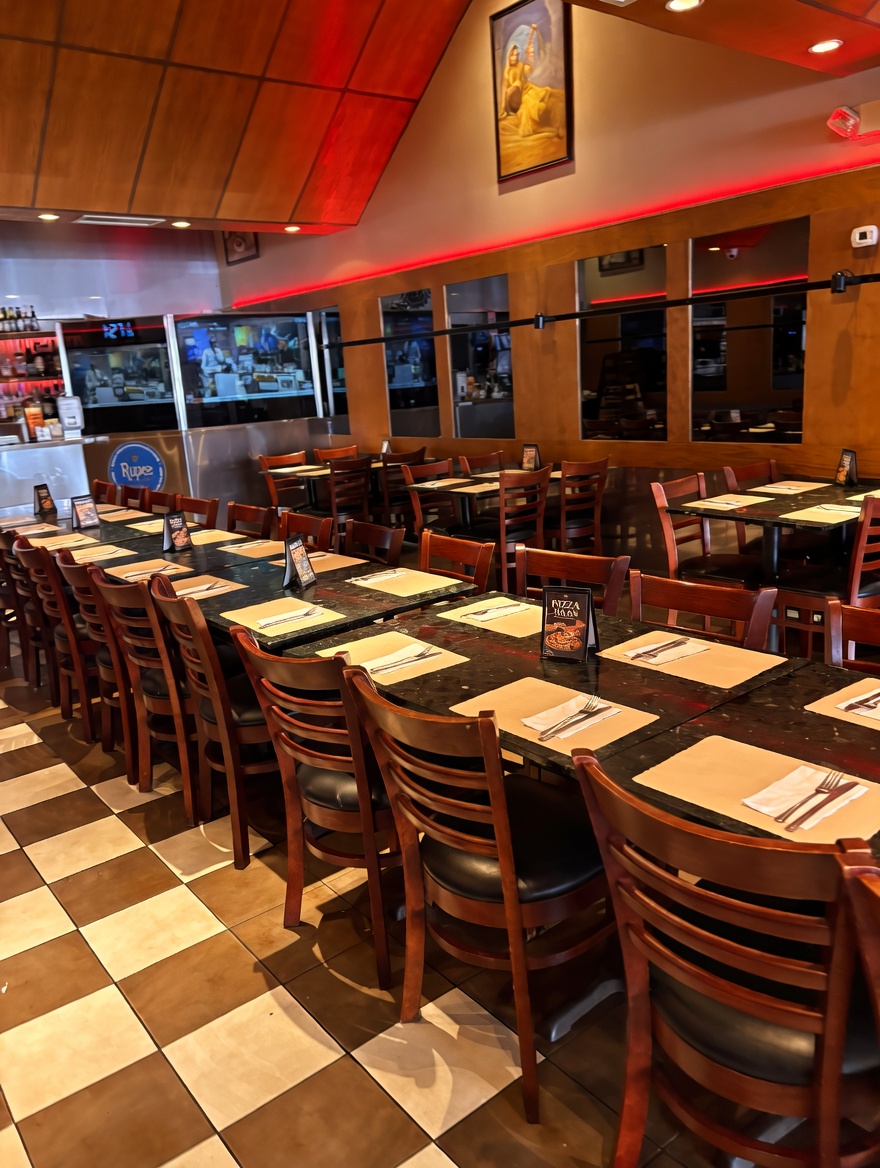 Spacious main dining room at New Delhi Indian Restaurant with long tables set with paper placemats and wooden chairs, checkered tile floor, red ceiling cove lighting, framed artwork, and visible kitchen area featuring the blue 'Rupee' sign