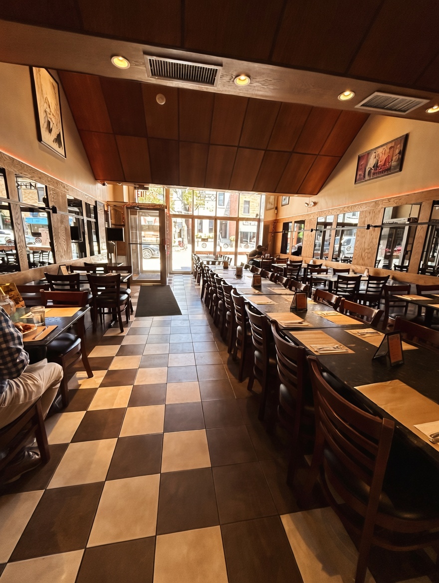 Wide view looking down the length of the dining room at New Delhi Indian Restaurant, showing many tables, wooden chairs, checkered tile flooring, wood-paneled ceiling, and large front windows overlooking the street