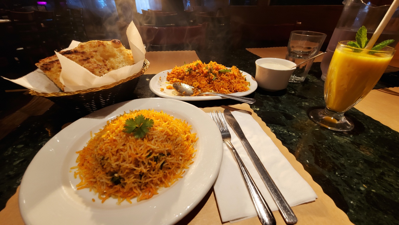 Table setting at New Delhi Indian Restaurant featuring aromatic biryani, a basket of fresh garlic naan, raita, and a refreshing mango lassi